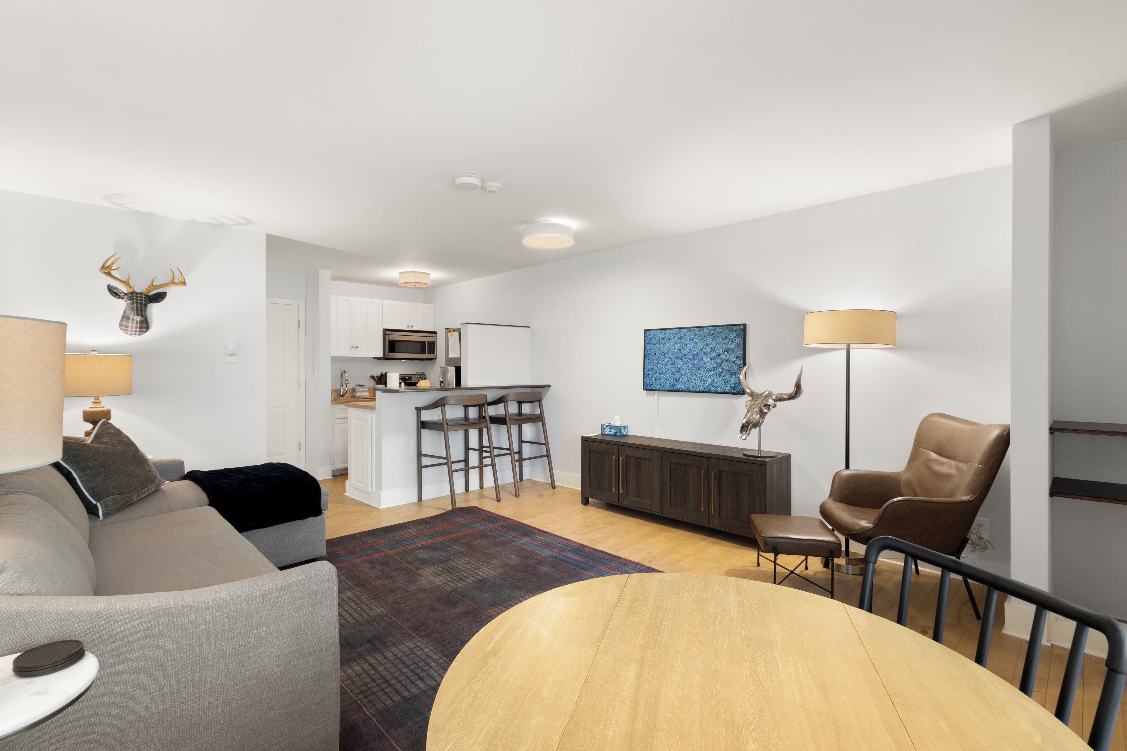 A modern living room with a grey sofa, a round wooden coffee table, a brown leather chair, and a kitchen in the background.