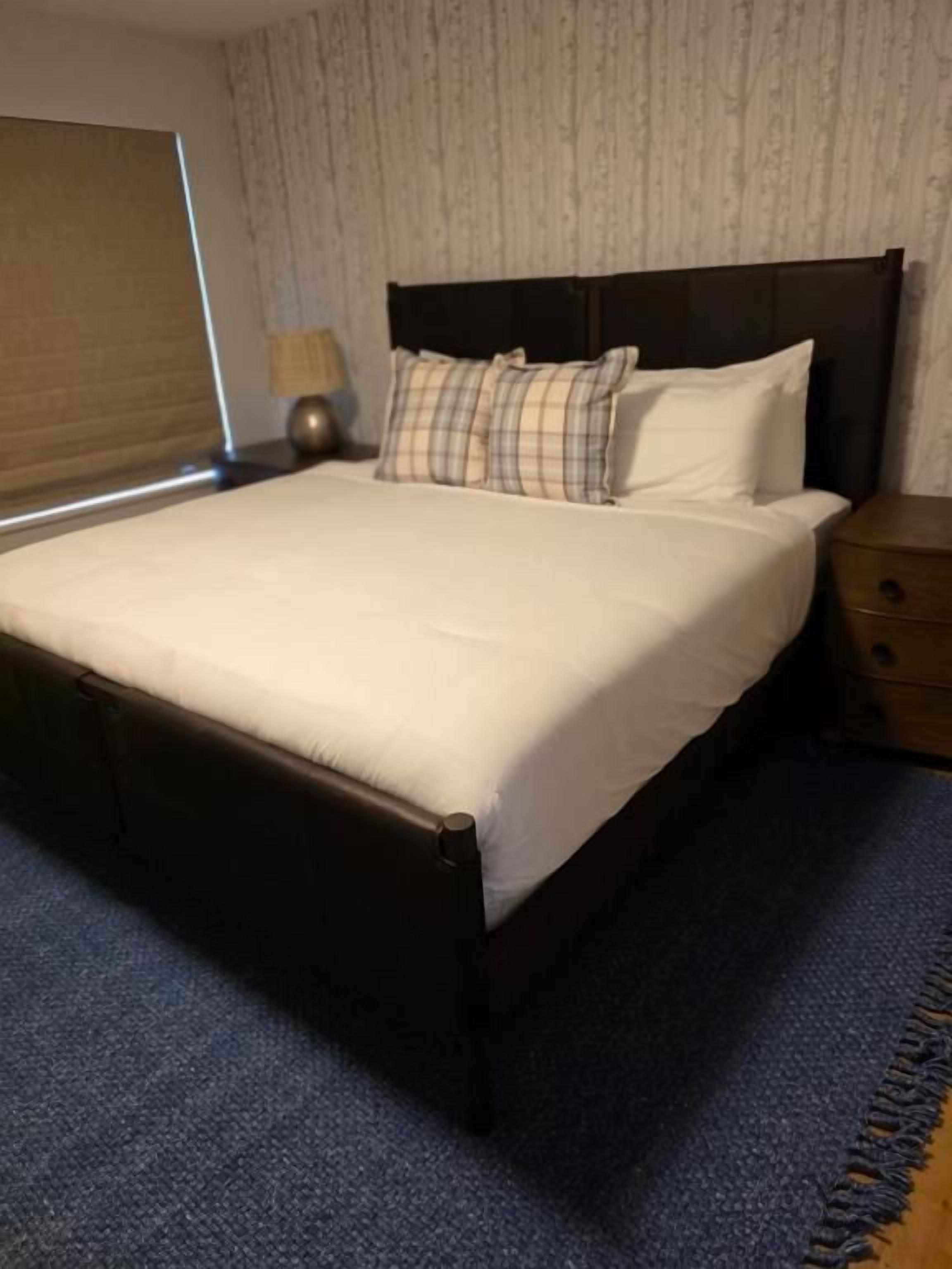A hotel room with a large bed, a nightstand, a lamp, and a window with blinds.