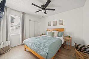 Ein Schlafzimmer mit einem Bett, einem Nachttisch, einem Deckenventilator, einem Fenster mit Jalousien und einer Tür.