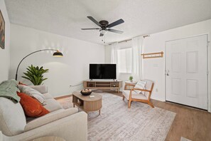 A living room with a ceiling fan, a flat-screen TV, a sofa, a coffee table, a chair, and a door. - Your Home Away From Home - Cozy Retreat (Bakersfield)