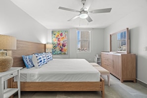 Ein Schlafzimmer mit Bett, Nachttischen, einem Schminktisch, einem Deckenventilator, einem Fenster mit Blick und einem farbenfrohen abstrakten Gemälde.