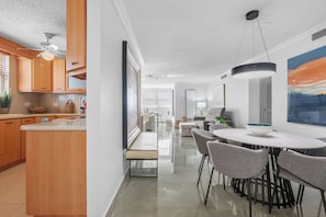A modern kitchen with wooden cabinets, a dining area with a round table and chairs, and a view into the living room. - Castillo Caribe by Otium (San Juan)