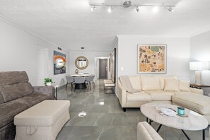 A modern living room with a grey sofa, a beige sofa, and a round coffee table. There is a dining area with a white table and chairs, and a hallway leading to another room. - Castillo Caribe by Otium (San Juan)