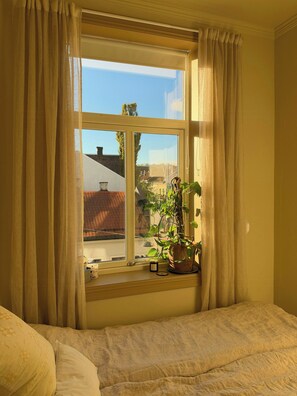 Ein Zimmer mit einem Bett, einem Vorhangfenster, einer Topfpflanze und einem kleinen Tisch mit Gegenständen.