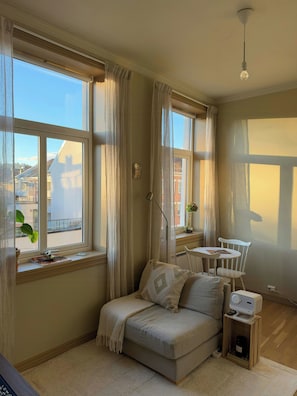 Ein gemütliches Zimmer mit einer Couch, einem kleinen Tisch und einem Stuhl. Ein Fenster mit Vorhängen, eine Pflanze und ein Blick auf umliegende Gebäude.