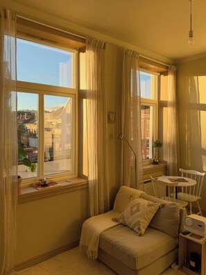 A cozy room with a sofa, a small table, and a chair. There are windows with curtains and a view of buildings outside. - A travel-curated flat in the heart of Grünerløkka (Oslo)
