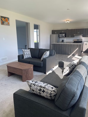 A modern living room with a grey sofa set, a wooden coffee table, and patterned cushions. A kitchen with grey cabinets and a built-in microwave is visible in the background. - Ottawa Getaway | 4BR Home + Free parking +WI-FI (Ottawa)