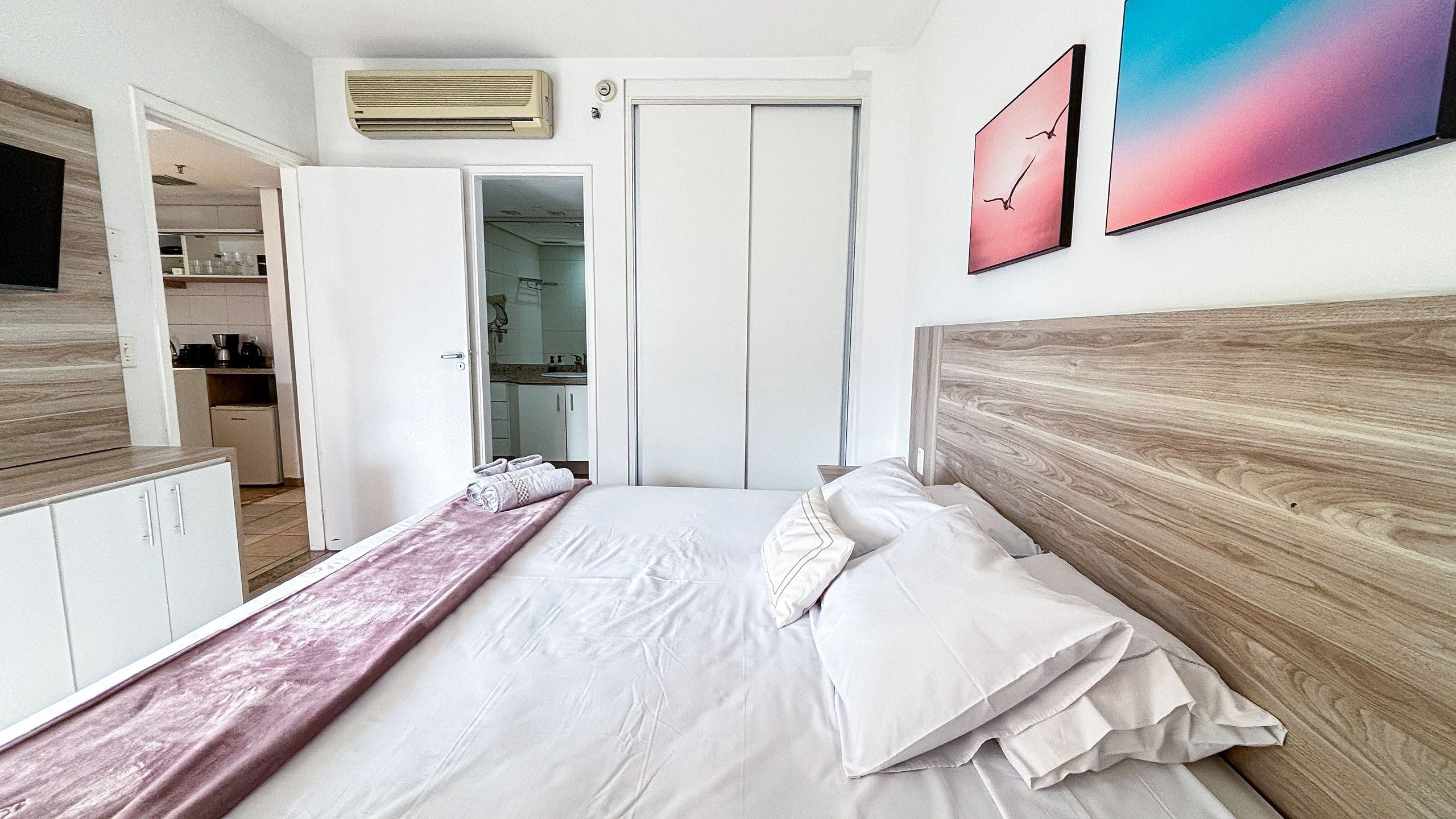 Un dormitorio con cama, cabecera de madera, mesita de noche, cómoda y baño visible a través de una puerta abierta.