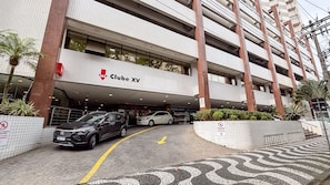 A modern building with a sign for 'Clube XV' and a parking area with two cars. - SA - Clube XV - Apto CXV1004 (Santos)