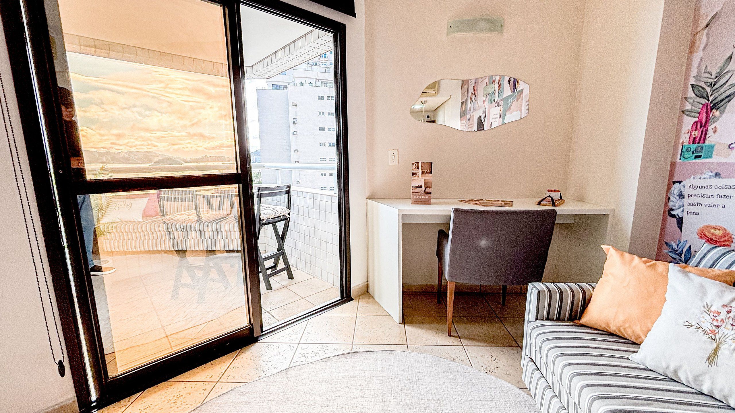 Quarto com porta de vidro deslizante para uma varanda, uma escrivaninha branca, um sofá listrado e uma pequena mesa com uma cadeira.