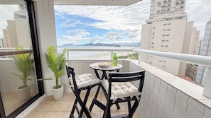 A balcony with two chairs and a table, overlooking a beach and cityscape. - SA - Clube XV - Apto CXV1004 (Santos)