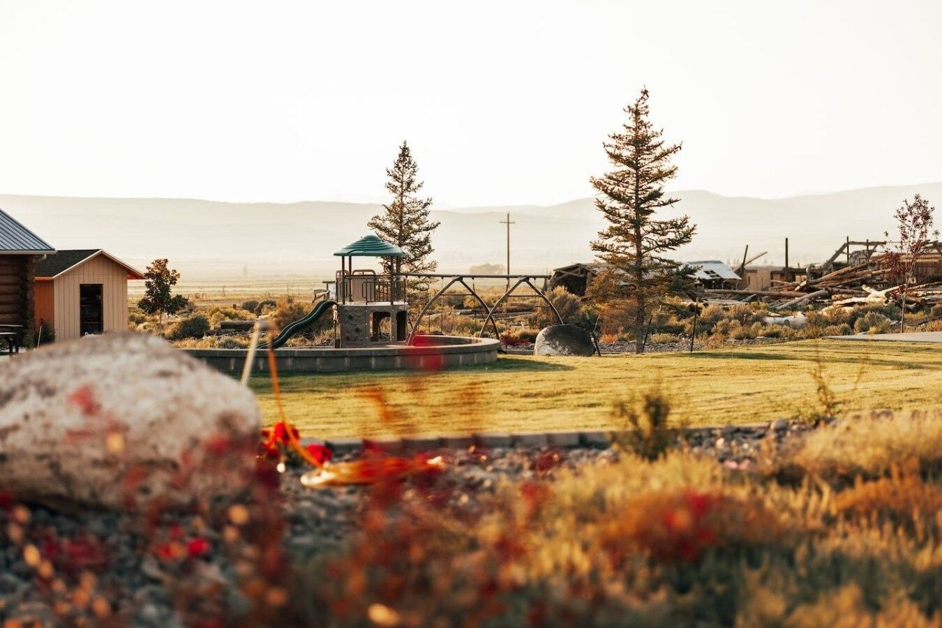 Un parque infantil con un tobogán y un pequeño refugio, rodeado de árboles y un campo cubierto de césped.