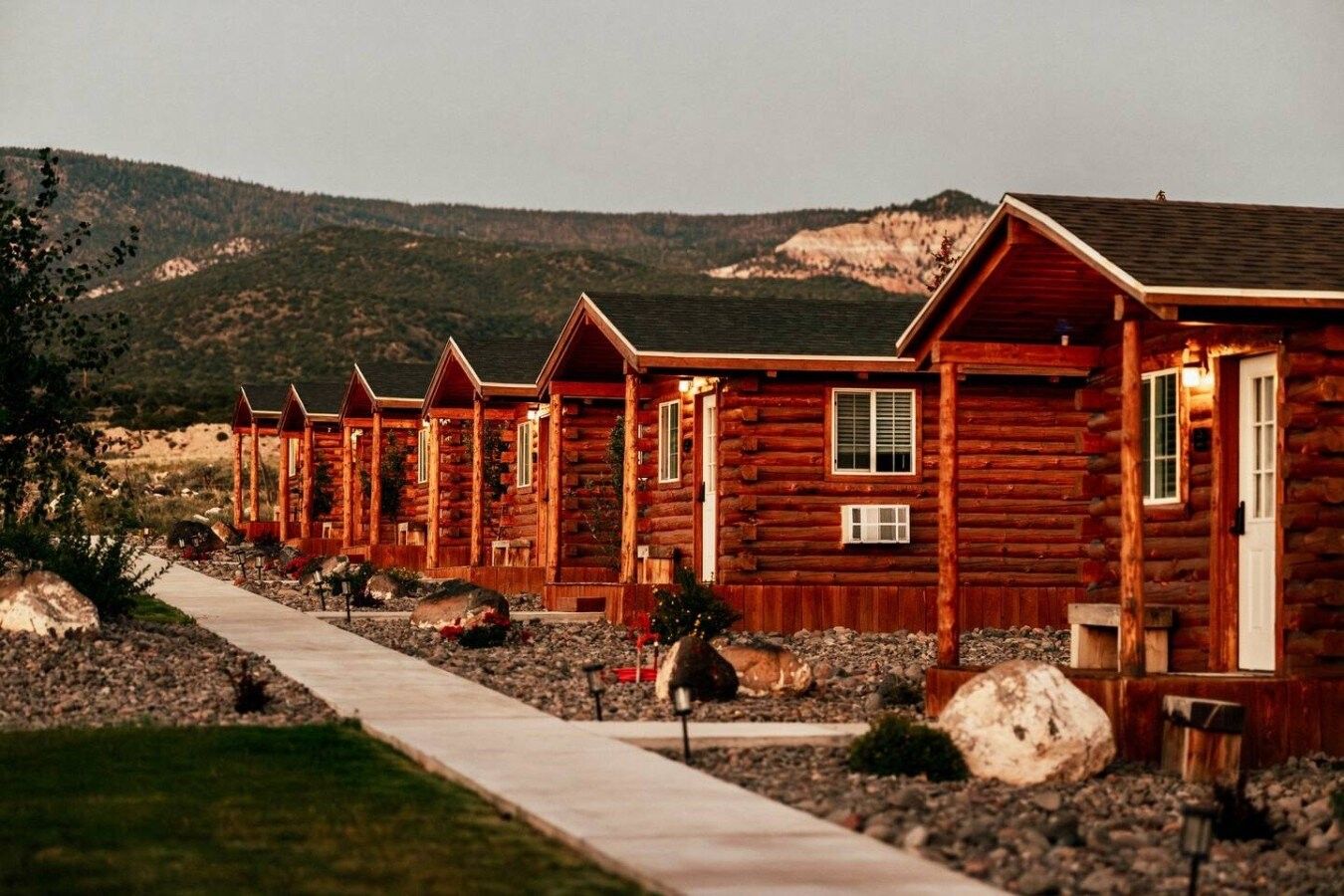 A row of log cabins with a gravel path leading to them, set against a mountainous backdrop.