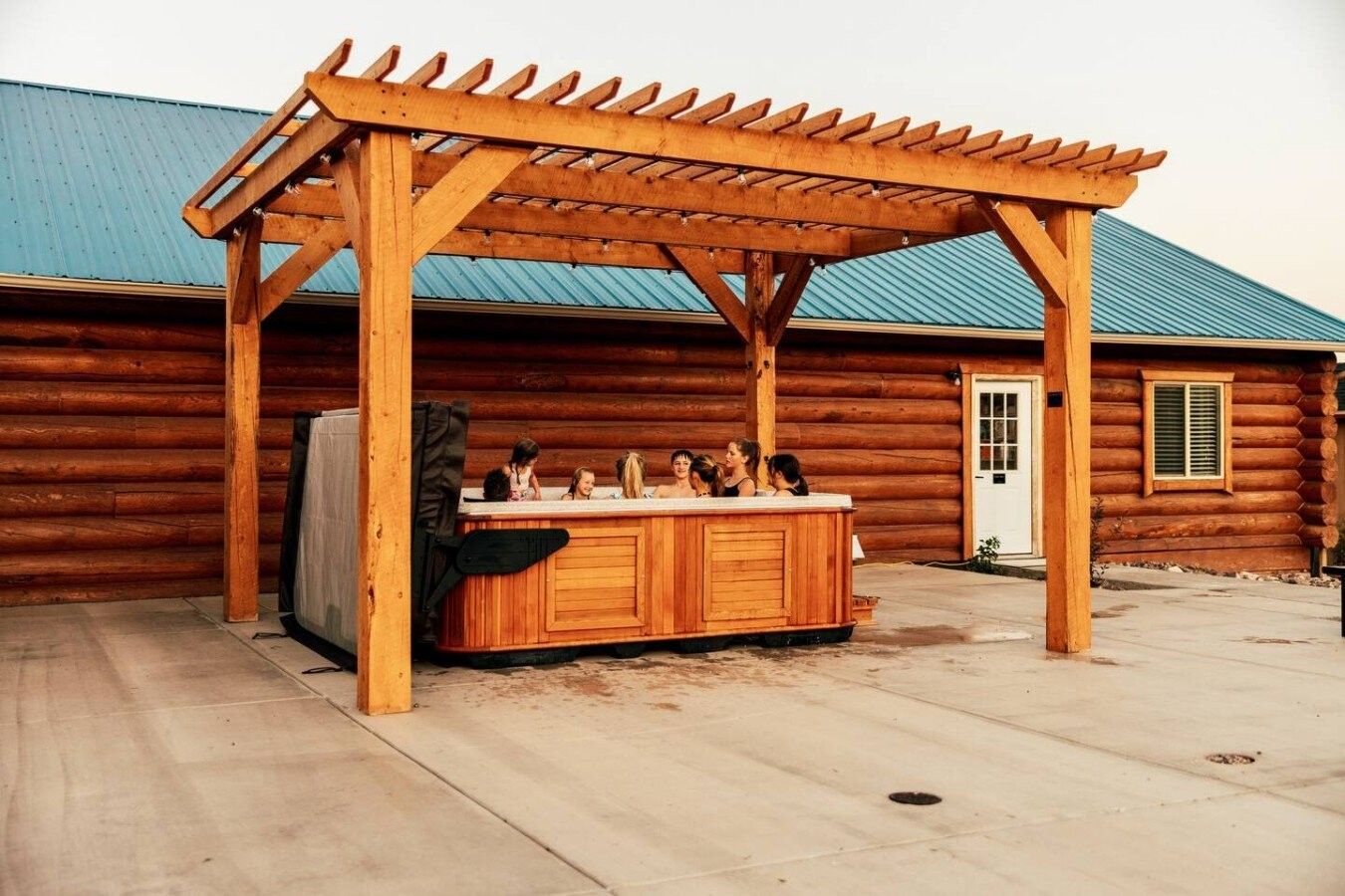 A wooden outdoor hot tub under a pergola with a log cabin in the background.