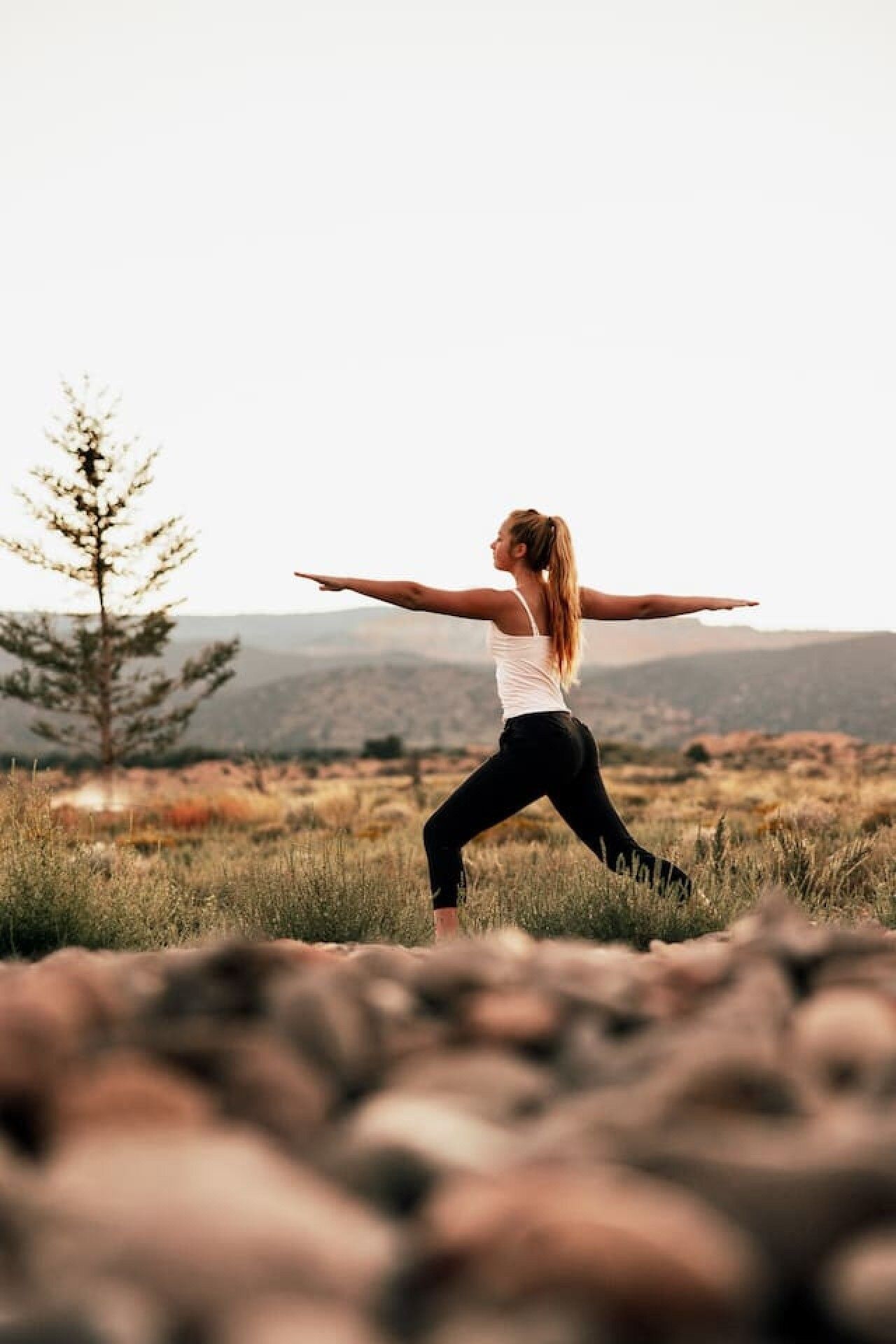 A person in a white tank top and black pants performing a yoga pose in an outdoor setting with a tree and hills in the background.