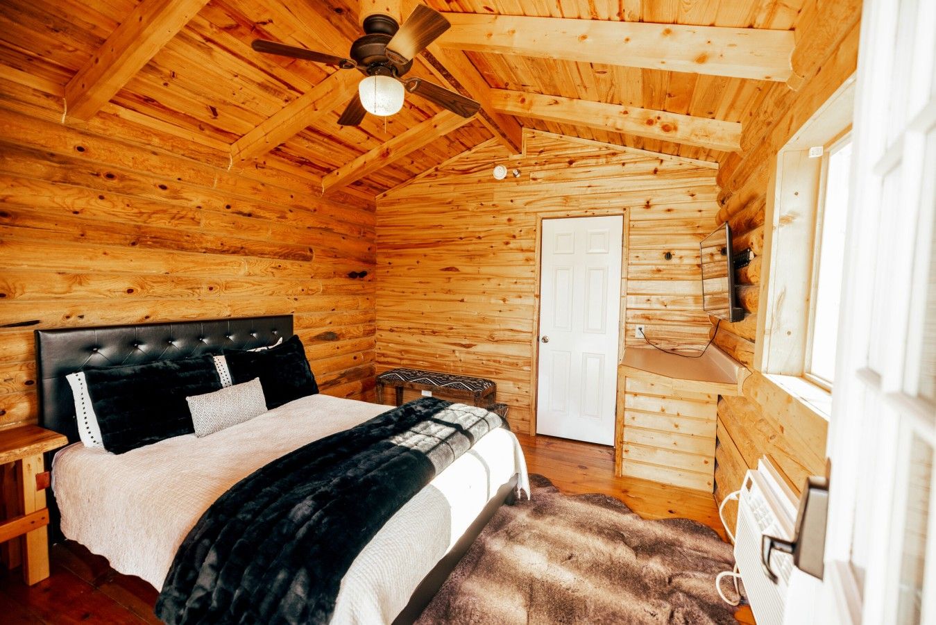 A wooden cabin bedroom with a bed, a ceiling fan, a door, and a window.