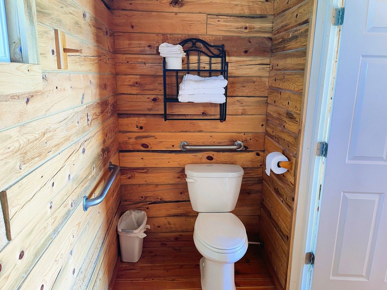 A compact wooden bathroom with a toilet, a handrail, a towel rack, and a trash bin.
