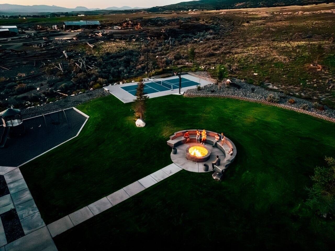An aerial view of a residential area with a circular outdoor seating area with a fire pit, a swimming pool, and a paved patio.