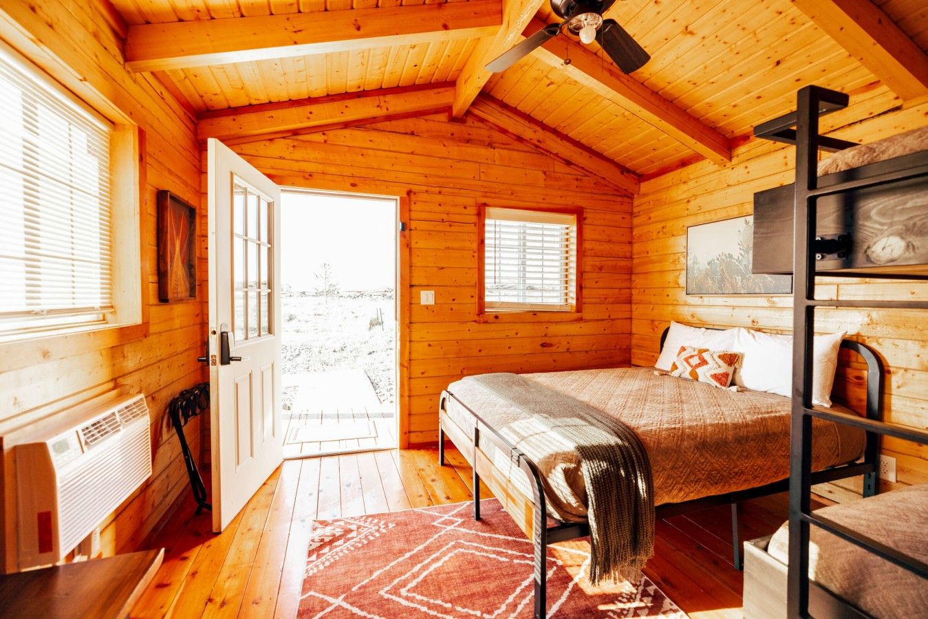 A cozy wooden cabin bedroom with a bunk bed, a heater, and a view through an open door.