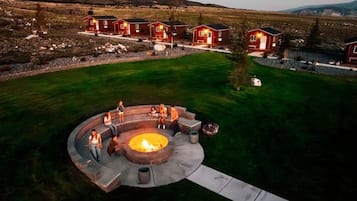 A circular outdoor seating area with a fire pit surrounded by stone seating, set against a backdrop of red cabins and a grassy field.