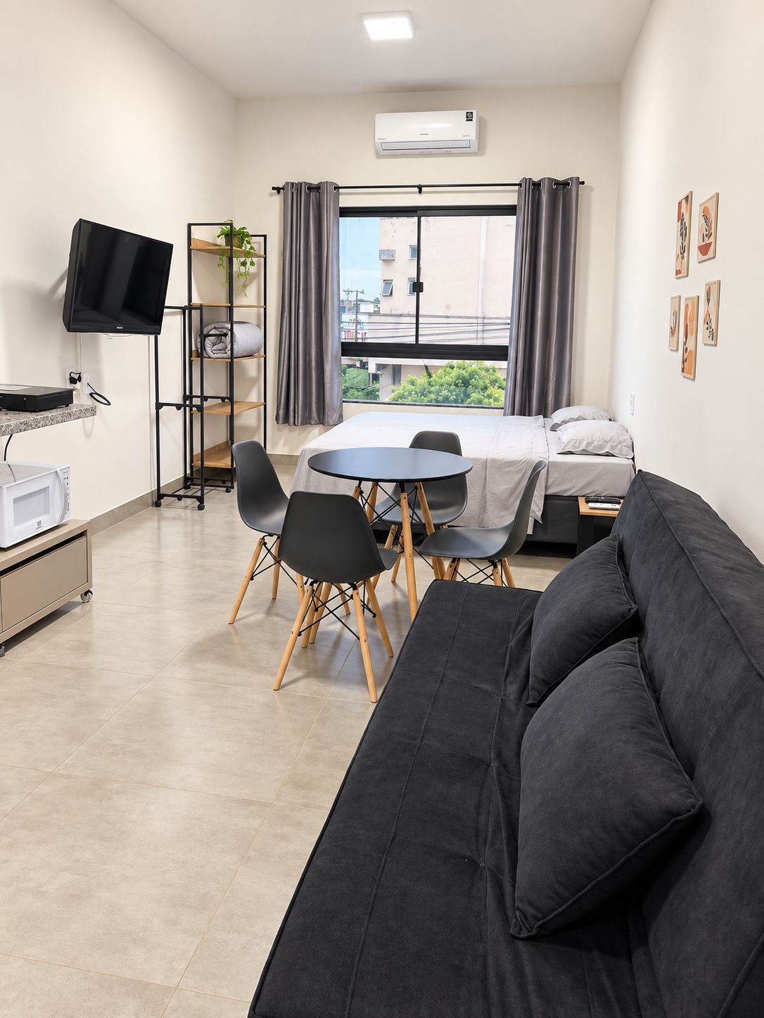 Una habitación de hotel compacta con cama, sofá, mesa de comedor con sillas, televisor montado en la pared y aire acondicionado.