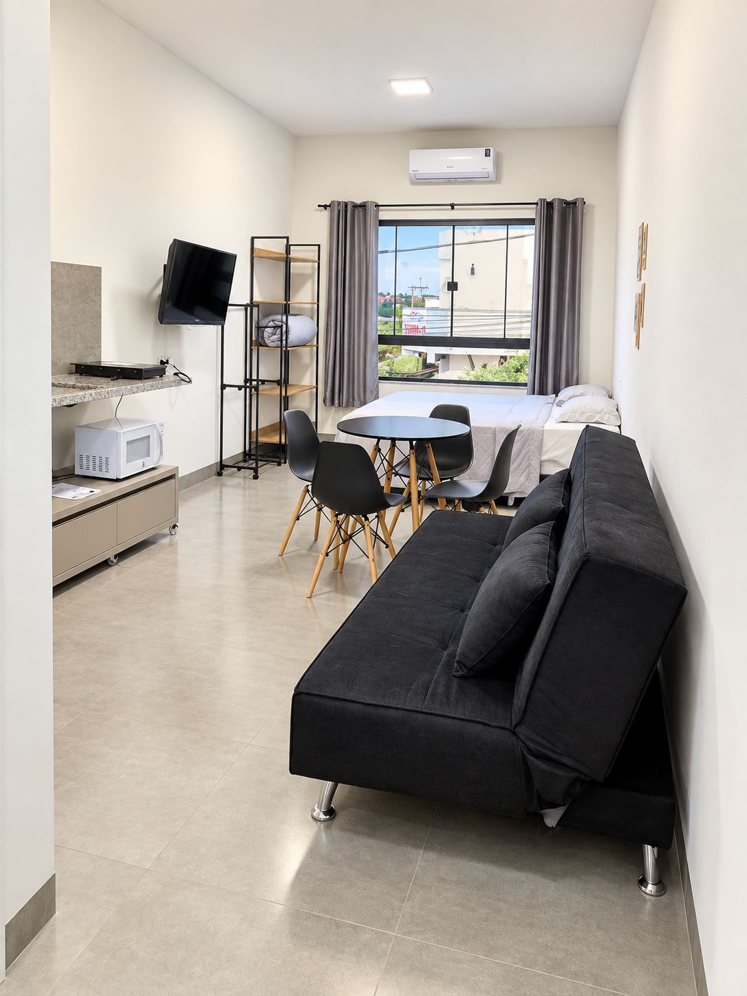 Een moderne kamer met een flatscreen televisie, een eettafel met stoelen, een bank, een kitchenette met een magnetron en een groot raam met gordijnen.