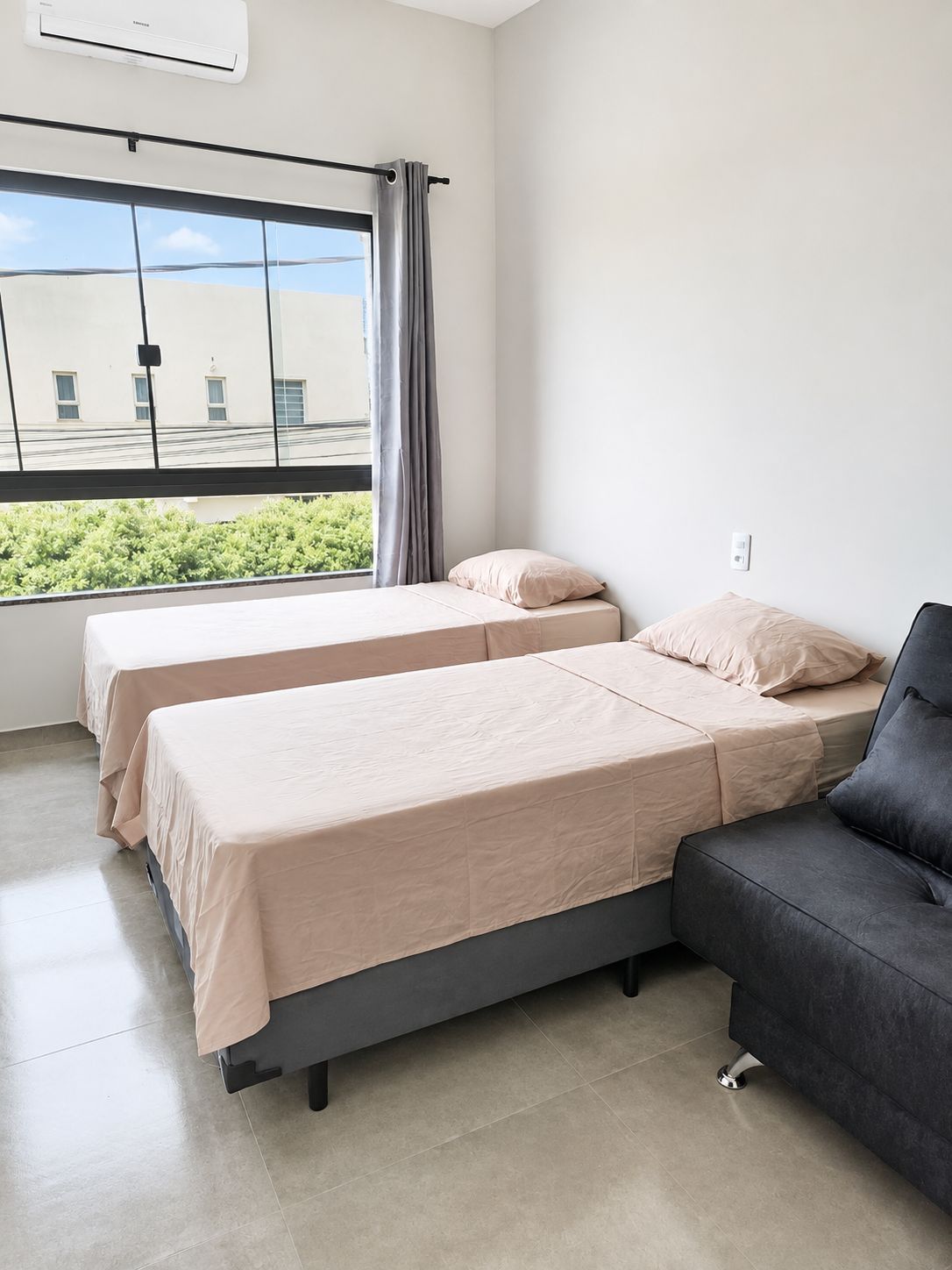 A hotel room with two beds, a sofa, a window with curtains, and a view of buildings and greenery.