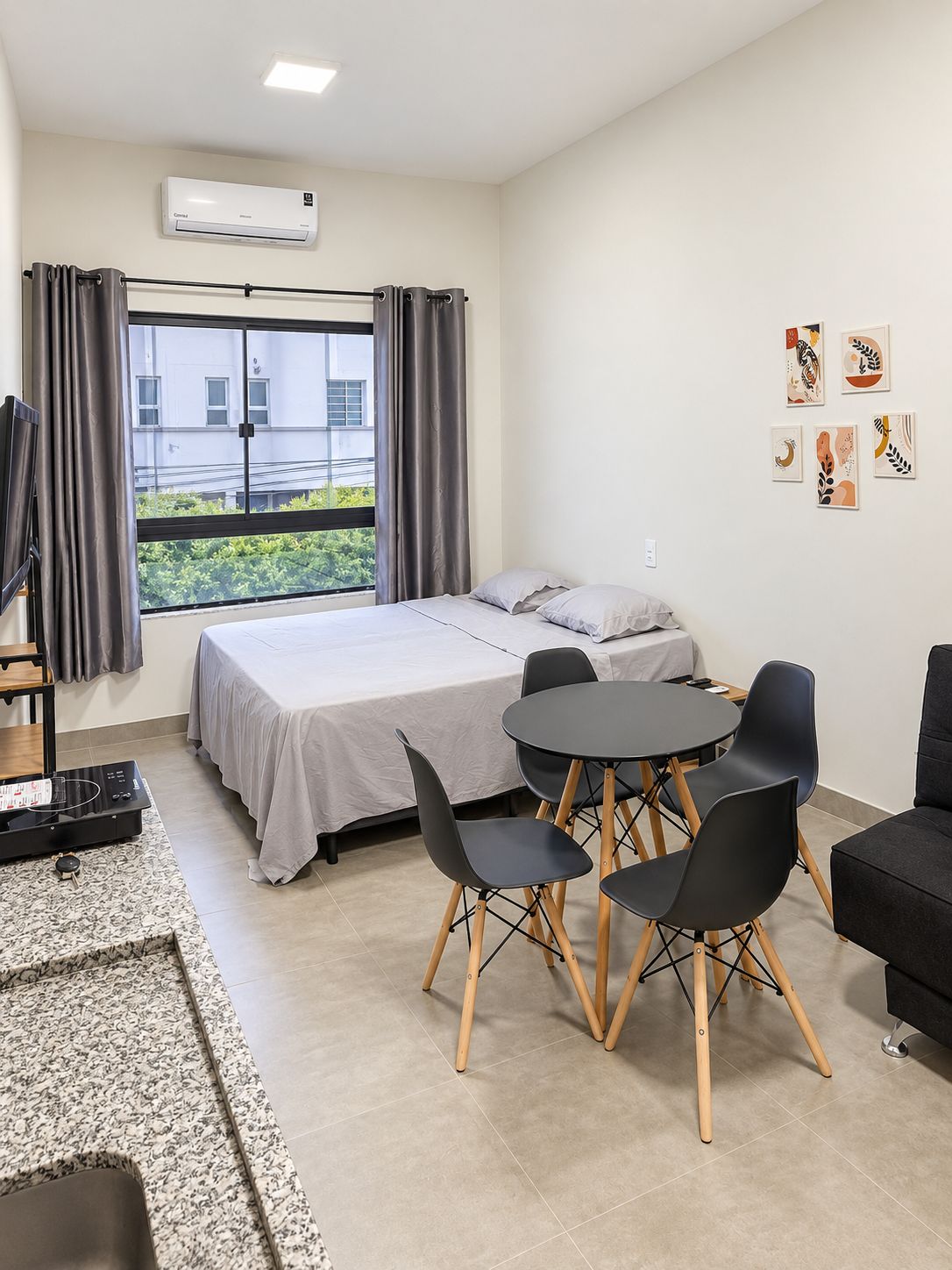A hotel room with a bed, a dining table with chairs, an air conditioning unit, and artwork on the wall.