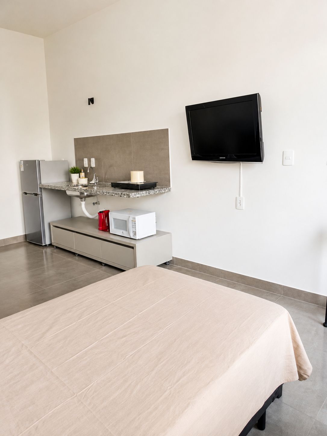Moderni hotellihuone, jossa on sänky, pieni keittiönurkkaus, taulutelevisio ja suuri peili.