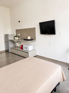 A modern hotel room with a bed, a kitchenette, a flat-screen TV, and a large mirror.