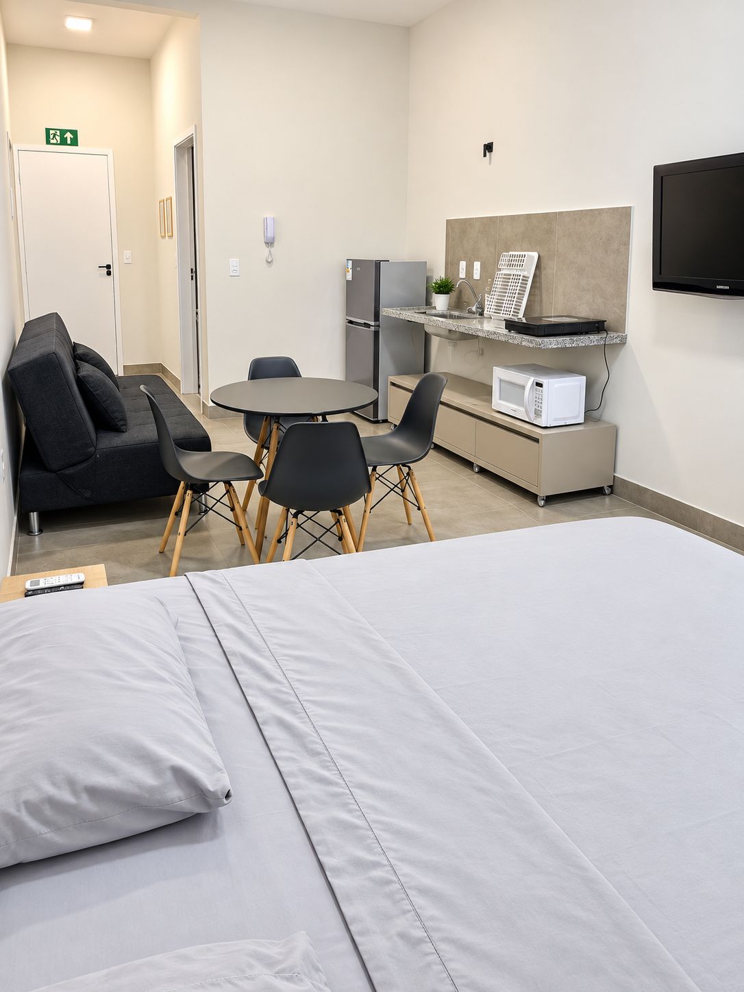 A hotel room with a bed, a small dining table with chairs, a kitchenette with a microwave, and a TV mounted on the wall.