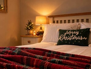 Un lit avec un coussin aux motifs de Noël, une lampe de chevet et un petit sapin de Noël posé sur une table de chevet.