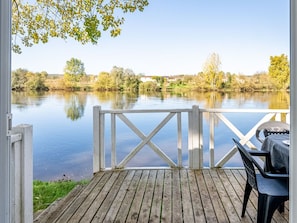 Eine Holzterrasse mit Grill, mit Blick auf einen ruhigen See, im Hintergrund Bäume und Häuser.