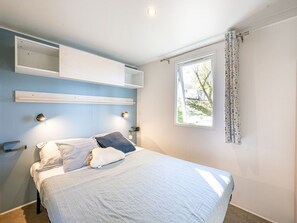 A bedroom with a bed, built-in shelving, a window with curtains, and a wall-mounted light fixture. - Mobile home with AC, pets welcome - 6 guests (Badefols-sur-Dordogne)