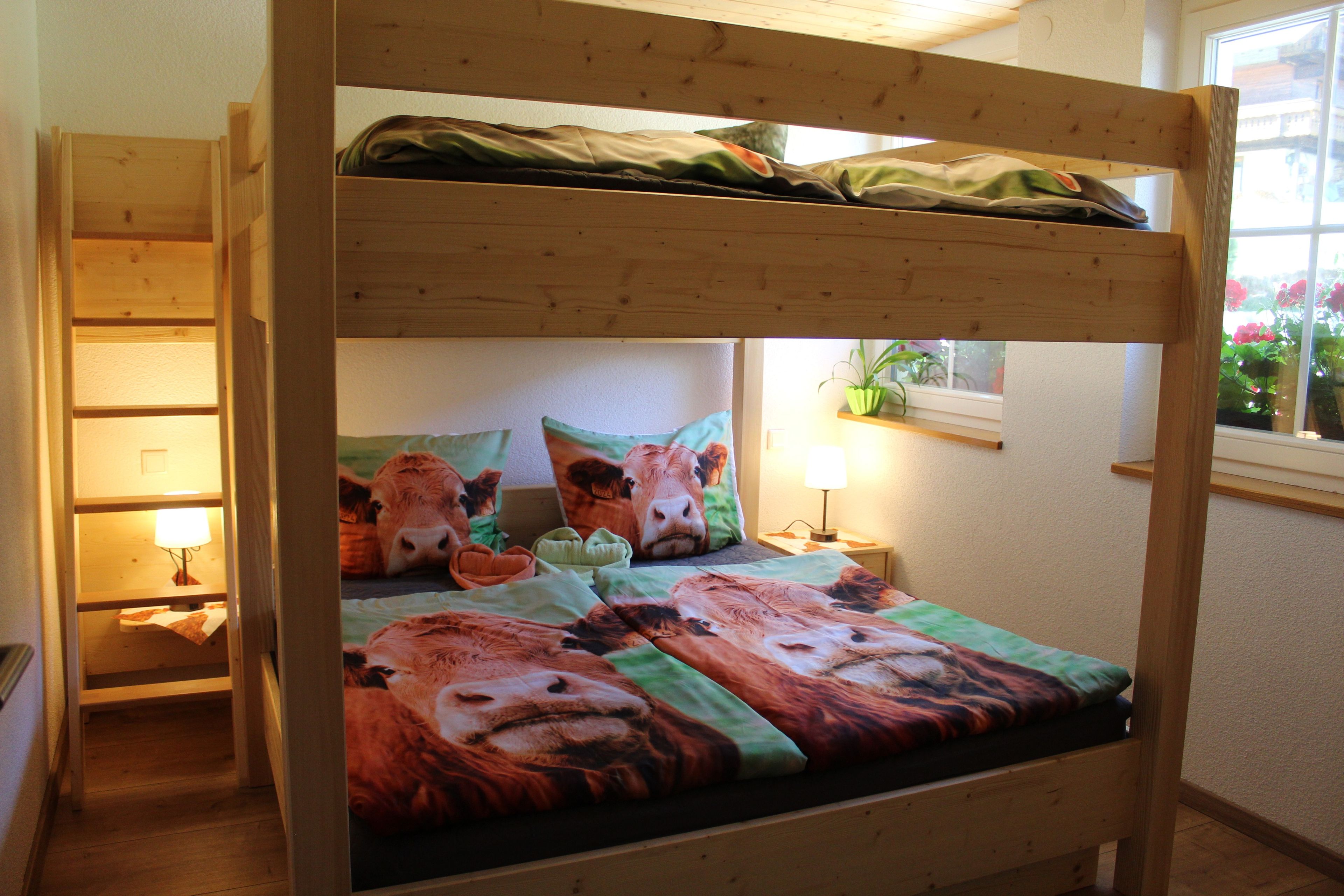 Una cama litera con colcha temática de vaca, una lámpara de noche y una ventana con flores.