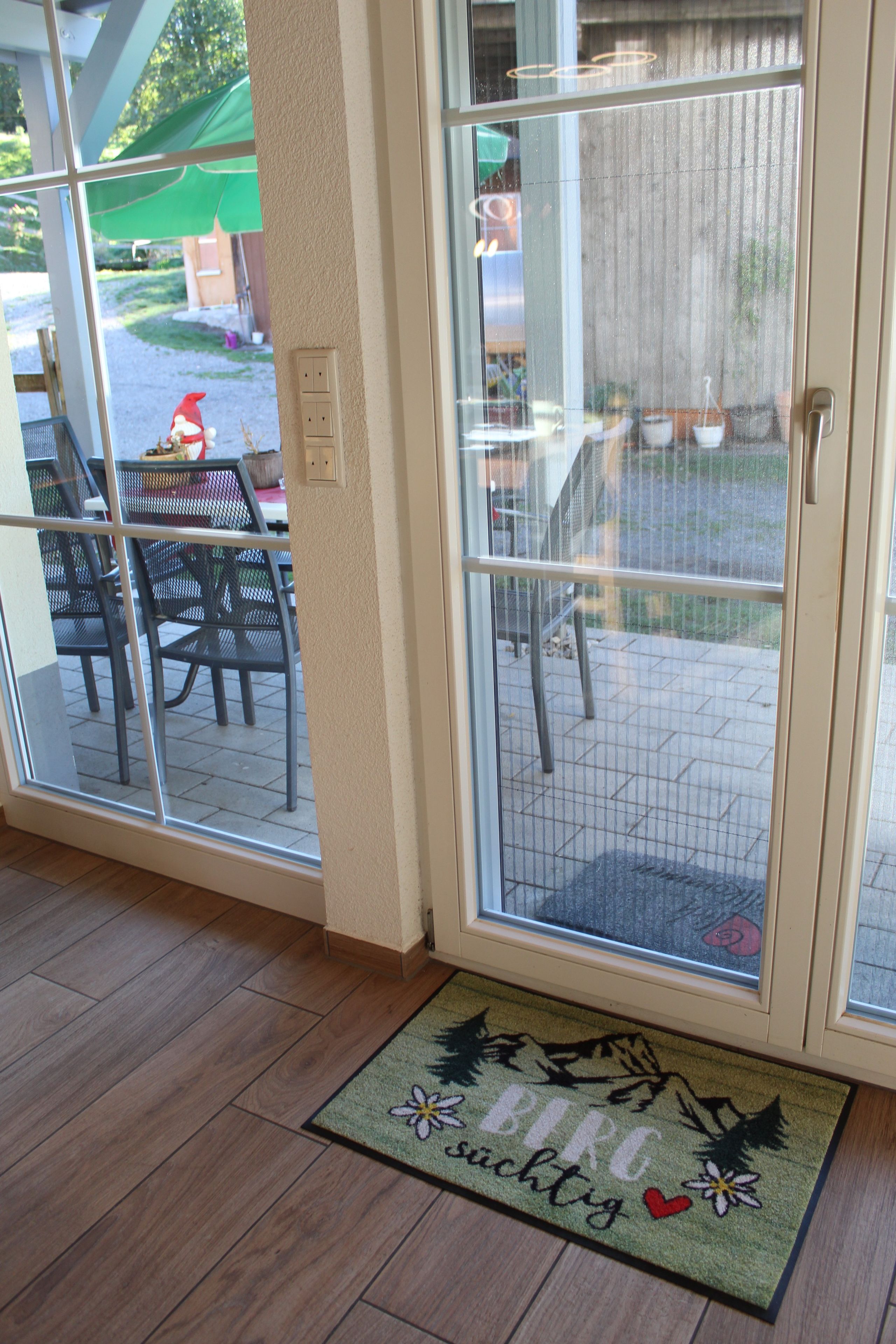 Puerta con un panel de vidrio, felpudo con texto y vista de mobiliario exterior.