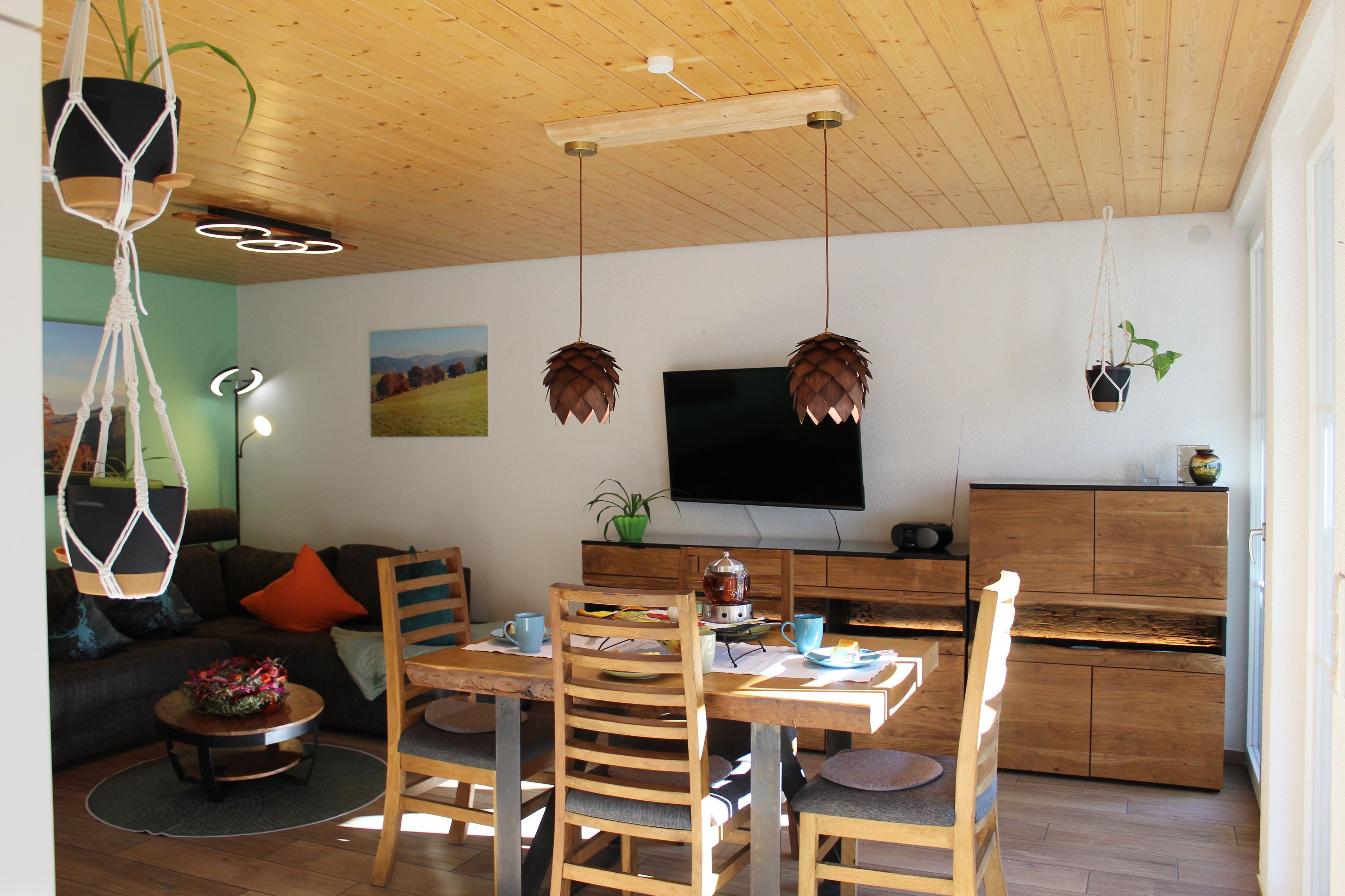 Um espaço de estar moderno com teto de madeira, área de jantar com mesa e cadeiras, sofá, televisão e cozinha com armários e bancada.