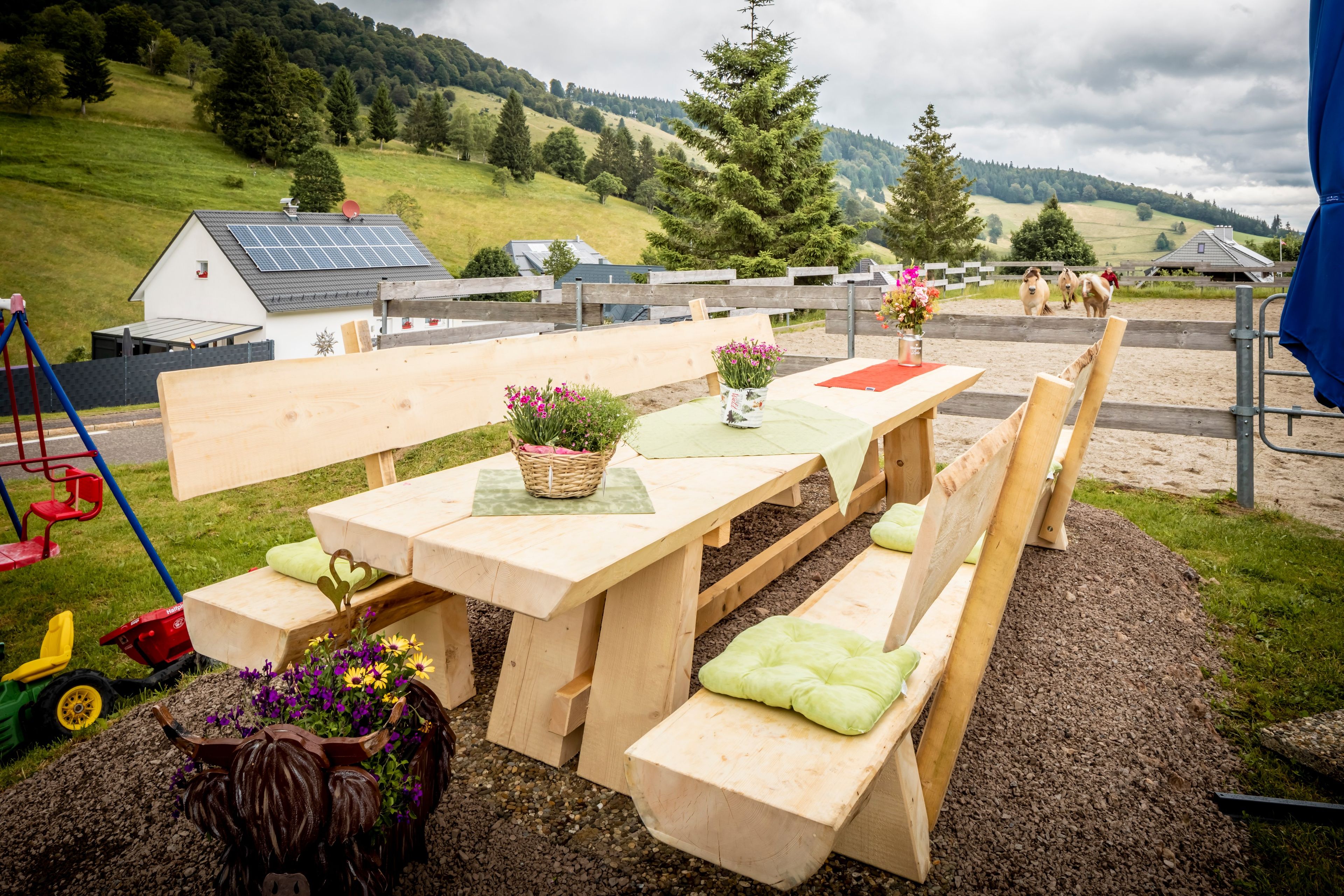 Ein Sitzbereich im Freien mit Holzbänken und Picknicktischen, umgeben von ländlicher Landschaft mit Hügeln und einem Haus im Hintergrund.