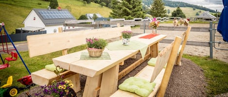 An outdoor seating area with wooden benches and picnic tables, surrounded by a rural landscape with hills and a house in the background.