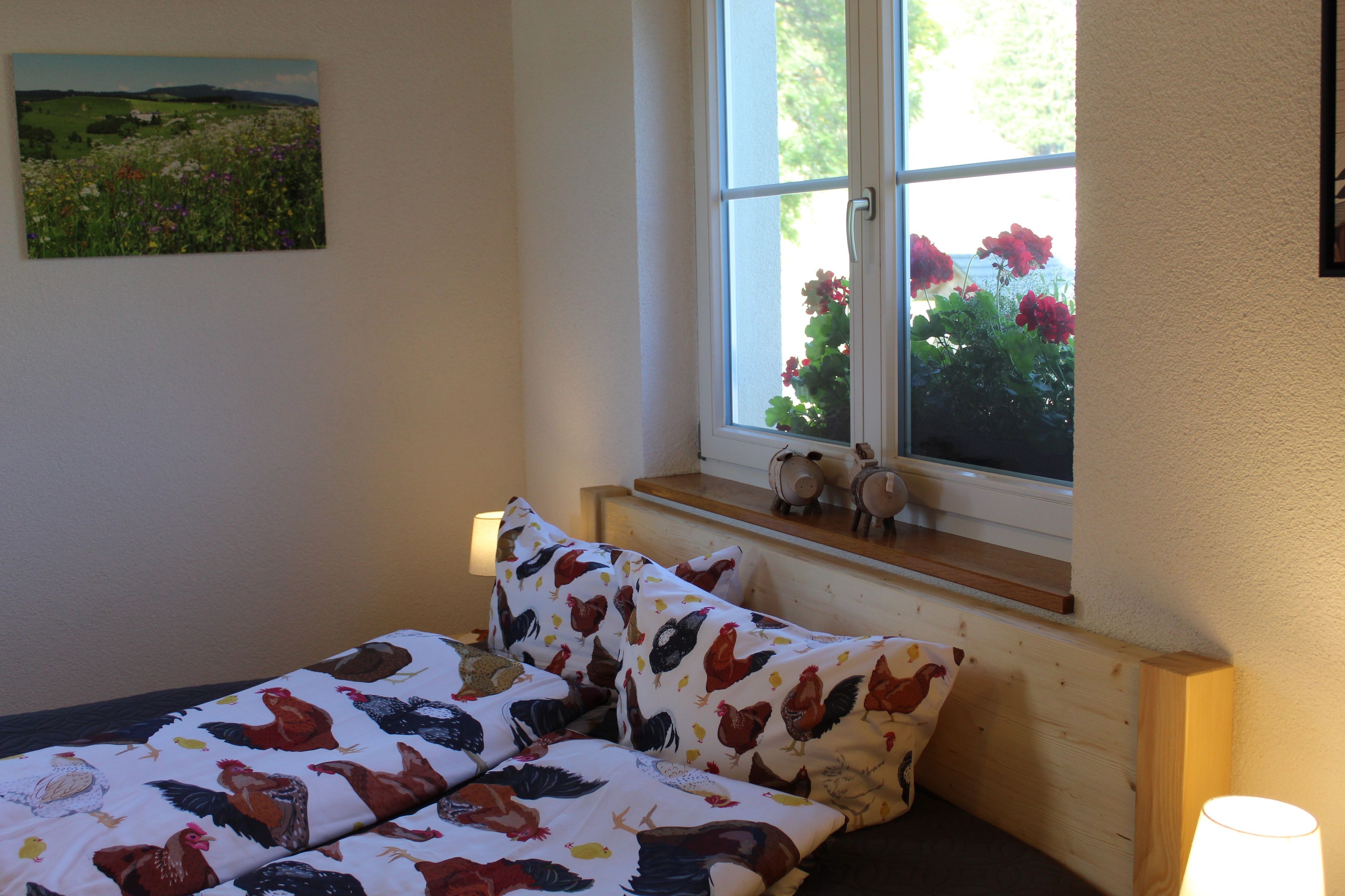A bedroom with a bed, a window with flowers, a lamp, and a painting on the wall.