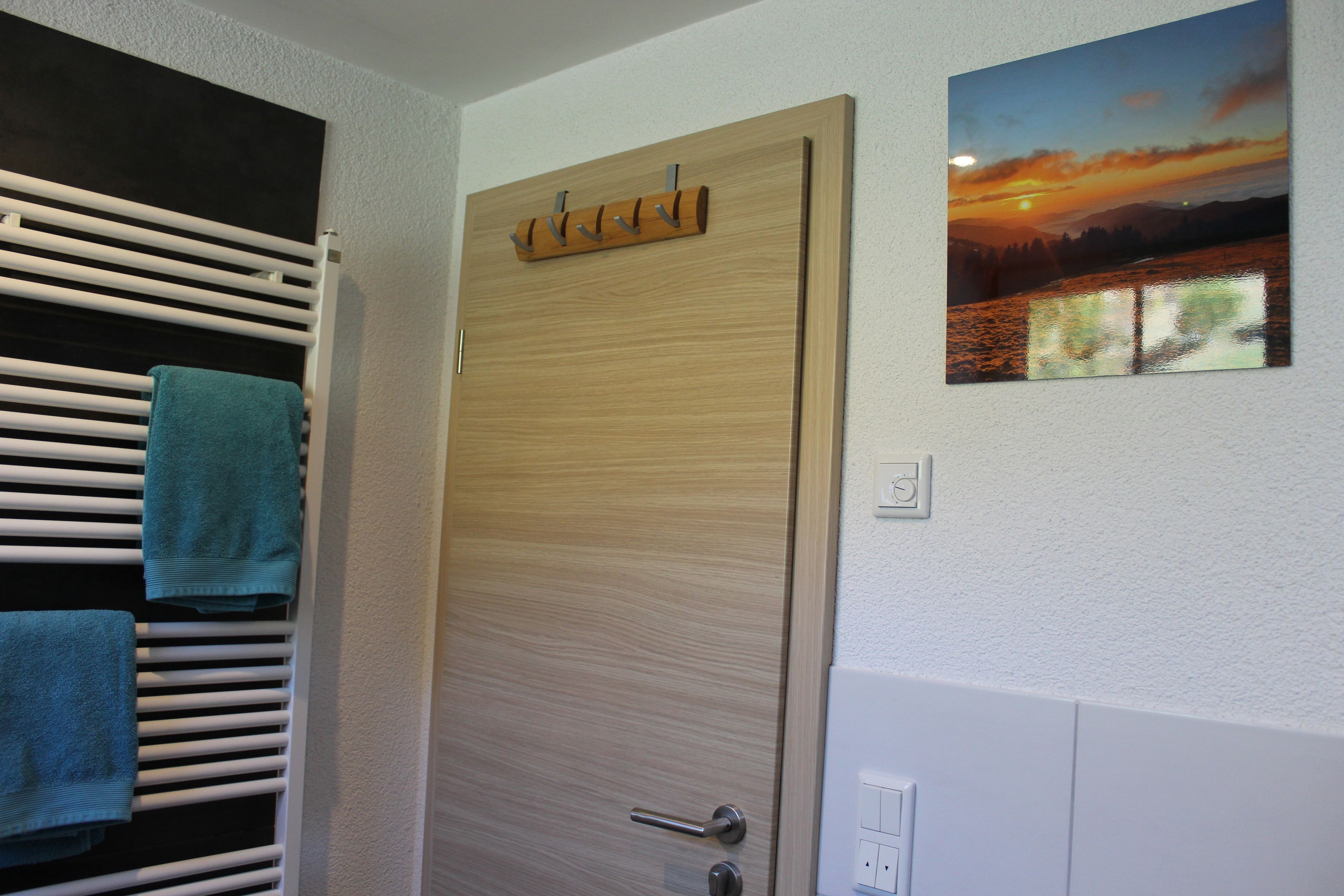 A room with a wooden door, a towel rack with towels, a wall-mounted picture, and a light switch.