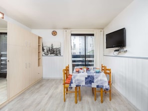 Ett kompakt vardagsrum med ett matbord för fyra personer, en tv som är monterad på väggen och utsikt över en byggnad genom ett fönster.
