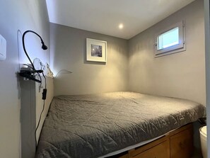 Un lit simple avec une couverture grise, une petite fenêtre laissant entrer la lumière du jour et un tableau encadré accroché au mur.