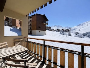 Un balcon avec une table et des chaises en bois, donnant sur un paysage montagneux enneigé parsemé de bâtiments.