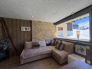 A cozy living room with a stone wall, a beige sectional sofa, a small table, and a window with a snowy view. - Luxury apartment, mountain view - 4 guests (Les Menuires)