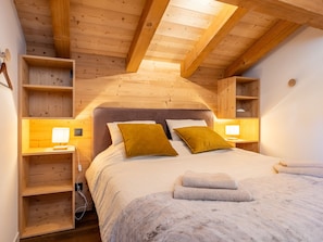 A bedroom with a wooden ceiling, a bed with pillows, bedside tables with lamps, and a shelf unit. - Half-chalet beautifully located, mountain view - 6 guests (La Clusaz)