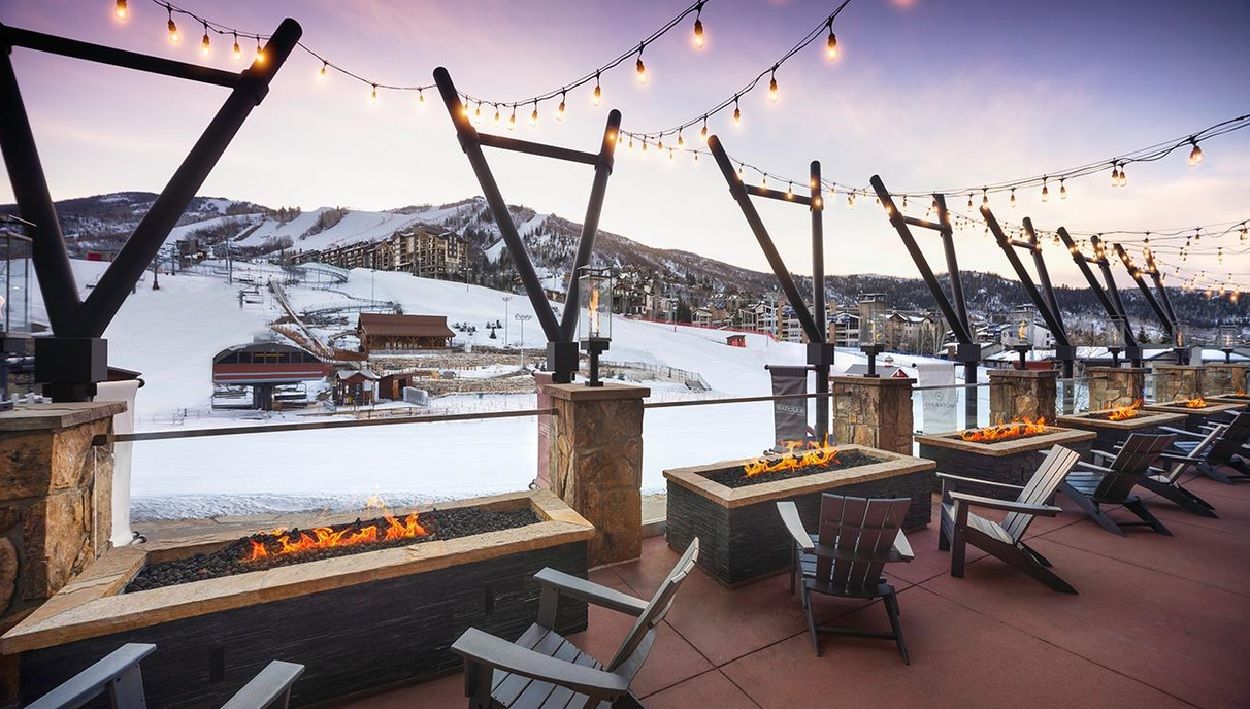 An outdoor seating area with fire pits and chairs, overlooking a snowy mountain landscape.