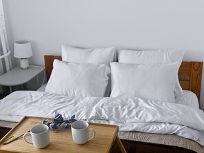 Ein ordentlich bezogenes Bett mit weißer Bettwäsche und Kissen, ein Nachttisch mit einer Lampe sowie ein Tablett mit zwei Tassen und einer Blume.