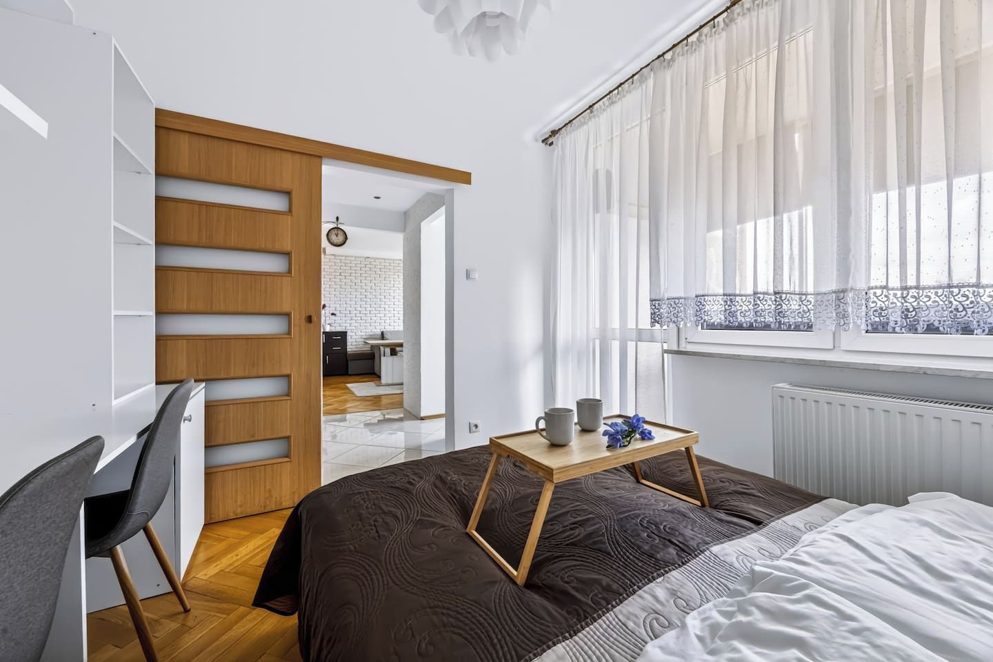 A bedroom with a bed, a desk, and a chair. There is a window with curtains and a view of a kitchen.
