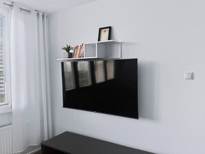 Uma sala de estar moderna com televisão de tela plana na parede, prateleira branca com objetos decorativos e janela com cortinas brancas.